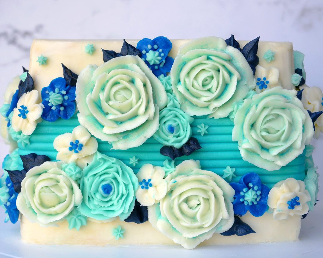 Wintery light blue and dark blue buttercream flowers decorate a white square buttercream cake with a blue textured stripe down the middle.